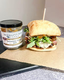 A sandwich with arugula, melted cheese, and caramelized onion on ciabatta rests on parchment beside a jar of Terrapin Ridge Farms Balsamic Onion Jam.
