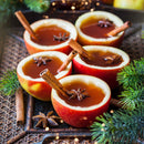 Five apple cups filled with spiced cider sit on a woven tray, each garnished with The Spice Lab Organic Cinnamon Sticks and star anise, surrounded by pine branches and festive lights.