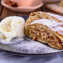 A slice of apple strudel dusted with powdered sugar sits on a plate next to a scoop of vanilla ice cream, the warm aroma hinting at a touch of The Spice Lab Apple Pie Spice Blend. In the blurred background, cinnamon sticks and a dish evoke the comforting essence of seasonal spice from The Spice Lab.