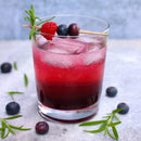 A glass of iced purple-red drink garnished with a rosemary sprig, a raspberry, and two blueberries on a skewer sits atop a gray surface, accented by scattered berries and rosemary. The inclusion of The Spice Lab Special Extra Bold Indian Black Peppercorns from The Spice Lab adds subtle warmth and complexity to this refreshing presentation.