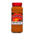 A 1 lb plastic jar of BBQ Bros Rubs Sweet Honey Cajun BBQ Rub, featuring a red lid and the BBQ Bros Rubs label, is displayed in front of a fiery background, highlighting its vibrant orange Cajun seasoning blend.