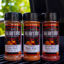 Three spice bottles from the BBQ Bros Rubs Western Style BBQ Rub Gift Set (3-Pack) – featuring California, Texas, and Kansas City Style Rubs – sit side by side on a grill, each adorned with flame motifs and set against a backdrop of softly blurred green foliage. This regional BBQ Bros Rubs set delivers authentic flavors from across the country, making it the perfect gift for any grilling enthusiast.