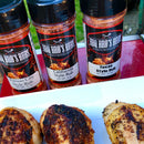 Three bottles from the BBQ Bros Rubs Western Style BBQ Rub Gift Set (3-Pack) by BBQ Bros Rubs are arranged behind a plate of grilled chicken breasts on an outdoor table.