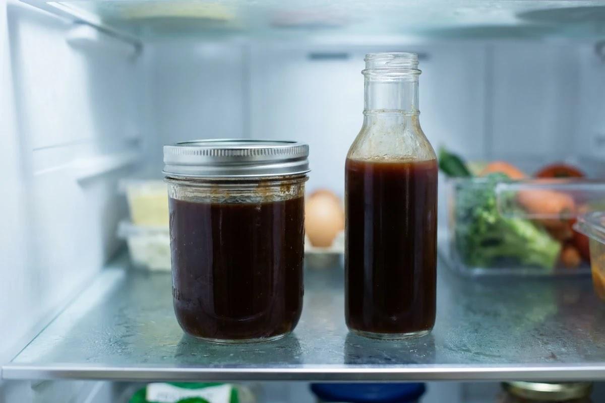 A mason jar and a glass bottle of dark BBQ sauce on a refrigerator shelf.