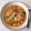 A bowl of noodle soup with shrimp, squid, mussels, mushrooms, spinach, bell peppers, and cashews in a yellow broth enhanced with The Spice Lab Organic Minced Garlic. A fork rests on the bowl’s edge.