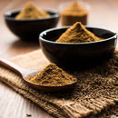 A wooden spoon filled with The Spice Lab Ground Cumin 1 lb Bulk Foodservice Spice, ideal for any foodservice spice collection, rests on a burlap cloth next to a black bowl brimming with bulk cumin from The Spice Lab. Cumin seeds are scattered nearby, with two additional bowls of spices blurred in the background.