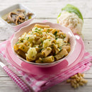 A bowl of rigatoni pasta with cauliflower, The Spice Lab Organic Pine Nuts, and herbs is served on a pink plate next to a small dish of marinated mushrooms. A fork and knife rest on a pink checked napkin beside the dish.