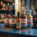 Five bottles from Bear & Burton’s Ultimate Sauce Bundle—including Worcestershire, Fireshire, Special Reserve Hot Sauce, and two Breakfast Sauces—are arranged on a rustic kitchen counter with spices, perfect for gourmet steaks or any meal.
