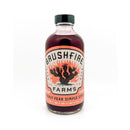 A glass bottle of Brushfire Farms Prickly Pear Simple Syrup, crafted from hand-harvested South Texas prickly pear, features a pink label with a cactus and flames design, displayed on a white background.