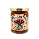 A glass jar of Brushfire Farms Peach Pepper Jam features a gold lid and a label adorned with a fiery cactus illustration, highlighting its handmade-in-Texas origin. This award-winning jam from Brushfire Farms is celebrated for its signature Hill Country Heat and has been recognized by the Good Food Awards.