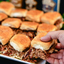 Hand holding a pulled pork slider with a tray of similar sliders in the background with big poppas grannys bbq sauce