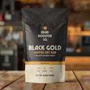 A black and gold Dead Rooster Co. Black Gold Coffee Dry Rub sits on a wooden surface against a blurred kitchen background. This bold BBQ rub for beef, chicken, and pork is an essential part of the Dead Rooster Co Variety Pack—featuring 3 signature BBQ rubs in one money-saving bundle.