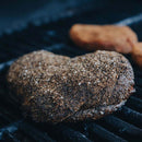 A thick, heavily seasoned steak—generously coated with flavors from the Dead Rooster Co. Variety Pack 3 Signature BBQ Rubs Bundle & Save—is grilling on a barbecue. Another piece of food is visible, blurred in the background.