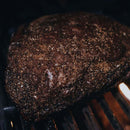 A close-up of a brisket covered in Dead Rooster Co. Black Gold Coffee Rub, sizzling as it smokes on the grill, with pronounced grill marks and a rich, dark crust forming to showcase the bold flavors of Dead Rooster Co.’s signature blend.