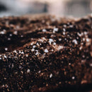 A close-up of a textured, dark brown surface sprinkled with coarse salt crystals, likely a well-seasoned piece of cooked meat enhanced with Dead Rooster Co Black Gold Coffee Rub from Dead Rooster Co. The shallow focus creates a blurred background.