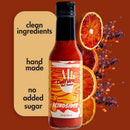 A bottle of Culture Hot Sauce Blindsided by Culture Hot Sauce stands against a dark brown background. To the left, a cream-colored text block reads clean ingredients, hand made, and no added sugar. On the right are blood orange slices and dried peppers, suggesting the bold flavors of this smoky citrus hot sauce.
