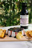 A wheel of baked brie topped with Terrapin Ridge Farms Blackberry Chipotle Sauce sits on a wooden board, cheese oozing beside toasted bread slices. A bottle of this Terrapin Ridge Farms sauce stands in the background, adding bold flavor to every bite.