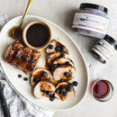 Sliced pork tenderloin topped with Terrapin Ridge Farms Blueberry Bourbon Pecan Jam is served with extra sauce, a golden spoon, two Terrapin Ridge Farms jam jars, and a glass of red liquid—perfect for charcuterie boards.