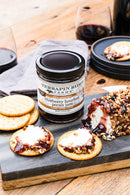 A jar of Terrapin Ridge Farms Blueberry Bourbon Pecan Jam Quart sits on a slate board with crackers, cheese, jam, and pecans—perfect for charcuterie boards. A nutty cheese log and glasses of red wine complete the scene.