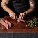 A person with tattooed arms uses the Steelport Knife Co. 6" Boning Knife to slice raw beef on a wooden cutting board, achieving precise meat cuts with fresh herbs and coarse salt nearby.