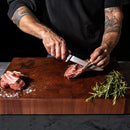 A person with tattooed arms is slicing raw lamb chops on a wooden cutting board using the Steelport Knife Co. 6" Boning Knife, handcrafted in the USA by Steelport Knife Co. Sprigs of rosemary and coarse salt rest nearby on the board, highlighting precise meat cutting. The background is dark.