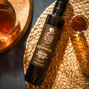 A bottle of Texas Hill Country Olive Co. Bourbon Reserve Balsamic Vinegar rests on a woven mat, accompanied by two glasses of amber-hued drinks and a partially visible wooden tray.