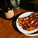 Four strips of glazed bacon rest on a wooden table beside a bottle of Under The Bark Syrup Co Bourbon Maple Syrup, sunlight highlighting the delicious pairing of smoky bacon and rich bourbon-infused maple syrup.