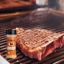 A thick steak with grill marks sizzles on the barbecue, steam rising, next to a pack of Hickman's BBQ Bovine Bonanza Beef Rub (Pack of 2).
