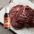 Two raw steaks sprinkled with coarse salt rest on a white cutting board. Next to the meat sits a bottle from the Hickman's BBQ Marinade Package by Hickman's BBQ. The scene is set against a rustic wooden surface.