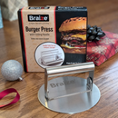 A Braize Smash Burger Press with Folding Handle & 50 Non-Stick Paper Sheets sits on a wooden table in front of its box. A silver ornament and wrapped gift nearby hint at festive, crispy smash burgers from Braize.