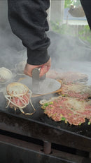 Using the Braize Smash Burger Press with Folding Handle & 50 Non-Stick Paper Sheets, a person presses a raw beef patty with onions onto a smoky outdoor griddle beside crispy-edged burgers and sliced onions.