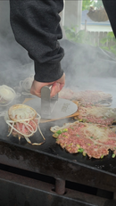 Using the Braize Smash Burger Press with Folding Handle & 50 Non-Stick Paper Sheets, a person presses a raw beef patty with onions onto a smoky outdoor griddle beside crispy-edged burgers and sliced onions.