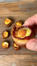 A hand holds a bacon-wrapped appetizer with a glazed center, while more appetizers sit on a wooden surface—perfect for dipping in Terrapin Ridge Farms Spicy Chipotle Garnishing Squeeze.