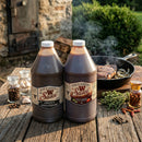 Two 64 oz bottles of Bear & Burton's W Sauce 2 Pack stand on an outdoor table by a cast iron skillet with steak and onions, surrounded by herbs and spices, with a rustic stone building in the background.