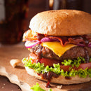 Close-up of a juicy cheeseburger with lettuce, tomato, red onion, bacon, melted cheese, and barbecue sauce in a toasted bun, topped with SALTOPIA Smoky Blended Salt: BURGER BUDDY by SALTOPIA Infused Sea Salts on a wooden surface.
