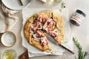 A savory galette topped with prosciutto and rosemary sits on parchment paper, partially sliced with a knife. Nearby are plates, a bowl of salt, charcuterie boards, white wine, and Terrapin Ridge Farms Strawberry and Fig Jam Quart.