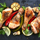 Four seasoned roasted chicken drumsticks with lemon slices, parsley, red and green chili peppers, roasted garlic halves, and aromatic The Spice Lab Special Extra Bold Indian Black Peppercorns arranged on a dark surface.