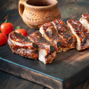 Juicy barbecued ribs sliced on a wooden cutting board, seasoned with The Spice Lab Organic Ground Black Pepper for an aromatic flavor, with a rustic clay pot and fresh cherry tomatoes in the background.