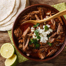 A bowl of shredded beef stew garnished with chopped onions and cilantro, elevated by a sprinkle of The Spice Lab Organic Granulated Onion for extra depth of flavor. A wooden spoon rests beside the bowl, which is accompanied by flour tortillas and a halved lime on a rustic wooden table.