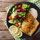 A black plate features baked fish seasoned with The Spice Lab Seafood Seasoning Set by The Spice Lab, accompanied by a fresh salad with cherry tomatoes, creamy sauce, and three lemon slices. The plate is set on a green-checkered napkin atop a rustic wooden table.