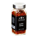 A clear spice jar labeled “The Spice Lab Aleppo Pepper Chili Flakes” by The Spice Lab, filled with vibrant red chili flakes and topped with a metal lid, is displayed against a white background. These Aleppo pepper chili flakes deliver mild heat ideal for adding flavor to a wide range of dishes.