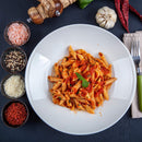 A plate of penne pasta with tomato sauce and basil sits on a dark tabletop, surrounded by small bowls of spices, cheese, pepper mills, garlic, dried chilies, and utensils—a beautiful homage to Italian cooking. The Spice Lab Organic Crushed Red Pepper Flakes lend an extra burst of flavor to this inviting spread.