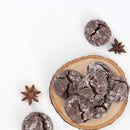 A wooden plate holding chocolate crinkle cookies is accompanied by The Spice Lab Ground Star Anise, imparting a sweet licorice-like flavor, with additional cookies placed on a white background.