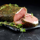 A close-up of herb-crusted cured meat with a slice cut and resting on a dark slate board, garnished with sprigs of The Spice Lab Whole Thyme by The Spice Lab, an aromatic seasoning that enhances the savory flavors.