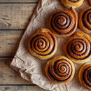 Six cinnamon rolls with golden-brown swirls are placed on crumpled parchment paper atop a rustic wooden surface. Freshly baked and neatly arranged, these pastries are infused with aromatic The Spice Lab Organic Ground Vietnamese Saigon Cinnamon for exceptional flavor.