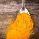 A metal spoon filled with The Spice Lab Golden Milk Turmeric Tea Superfood Premium Powder rests on a rustic wooden surface, with some of the bright orange superfood powder spilled beside it.