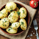A wooden bowl filled with boiled potatoes garnished with fresh dill and a sprinkle of The Spice Lab Organic Parsley Flakes, set on a burlap cloth. A fork, scattered salt, and a tomato rest nearby on the wooden table.