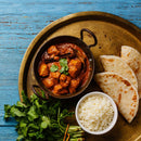 A bowl of chicken curry, subtly flavored with The Spice Lab Organic Ground Vietnamese Saigon Cinnamon and topped with fresh cilantro, is served alongside white rice and flatbread on a round brass tray atop a blue wooden table.