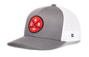 Introducing the Haka Hat Carolina Hockey Trucker Hat | Red CAR X Snapback: a gray and white trucker hat featuring a bold red circular patch on the front with two crossed hockey sticks, a puck, and the letters C, A, and R—celebrating Raleigh, North Carolina pride. The back is finished in classic white mesh for breathability and comfort.