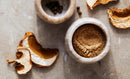 Two marble bowls—one filled with whole peppercorns and the other with World Spice’s Cascade Mushroom Mix—sit on a stone surface near several dried mushroom slices, making them perfect ingredients for adding an umami bomb to your soups and gravies.